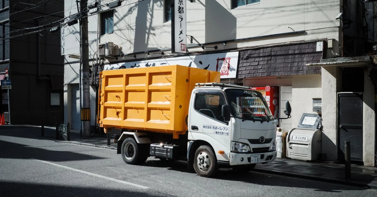 Camion benne jaune vif garé dans une rue urbaine à Kyoto, au Japon, avec des bâtiments environnants.
