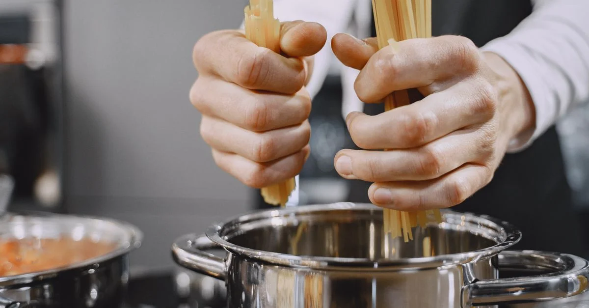 Gros plan sur les mains d'un chef ajoutant des pâtes dans une casserole bouillante dans une cuisine.