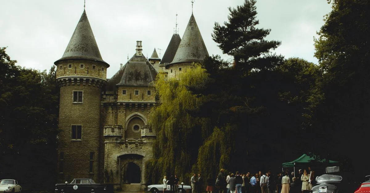 Un beau château en pierre avec des voitures anciennes et des personnes rassemblées en plein air par une journée nuageuse.