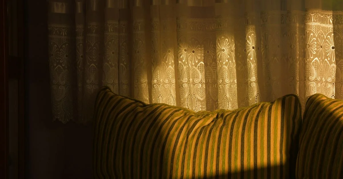 Des rideaux en dentelle illuminés par le soleil projettent des motifs sur un canapé rayé dans un cadre intérieur cosy.