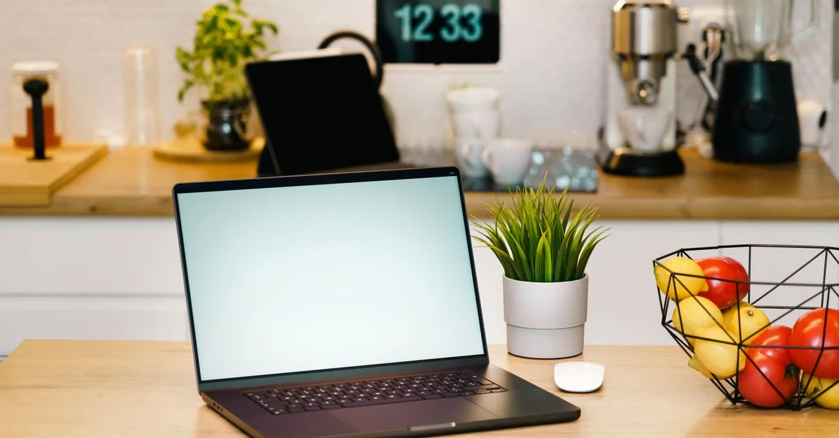 Un espace de travail de cuisine moderne comprenant un ordinateur portable, une machine à café et une corbeille de fruits, idéal pour le travail à distance.