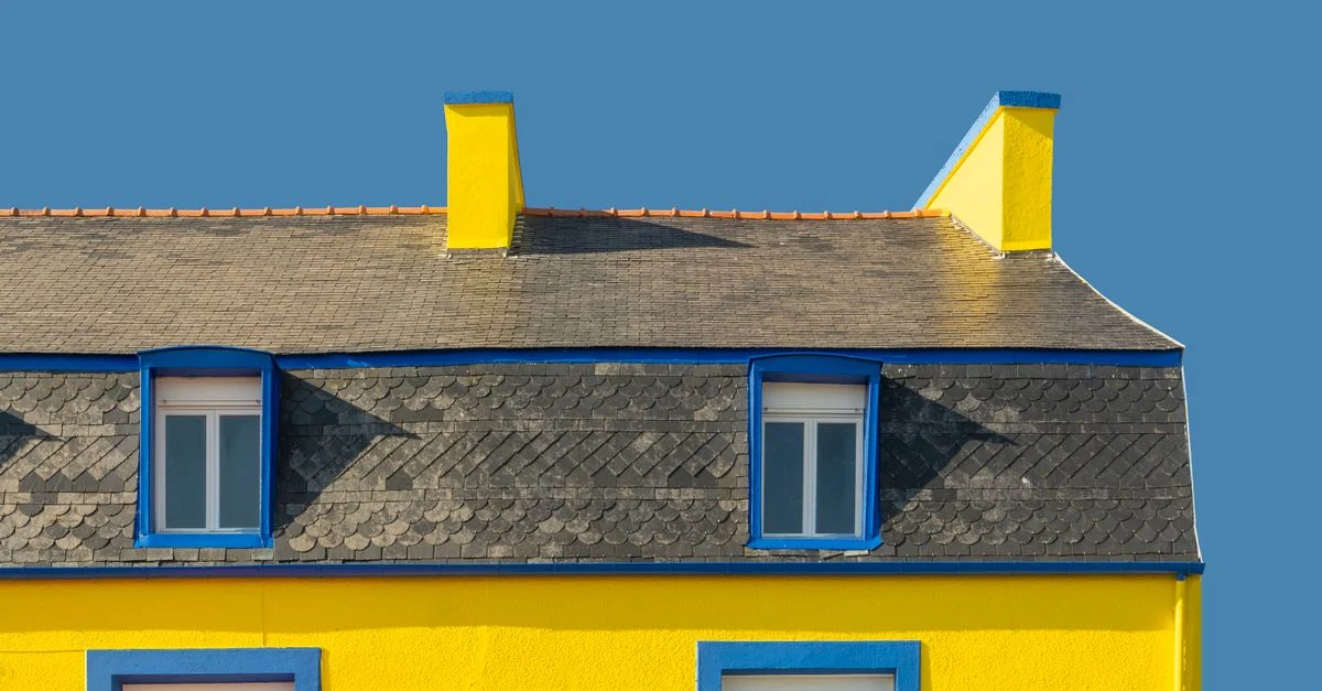 Une façade de maison jaune vif avec des accents bleus et des cheminées contre un ciel clair.