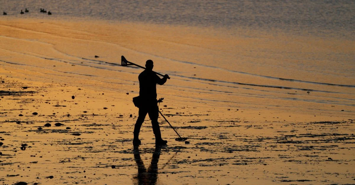 Silhouette d'un homme utilisant un détecteur de métaux sur une plage au coucher du soleil, créant une scène paisible et propice à la contemplation.