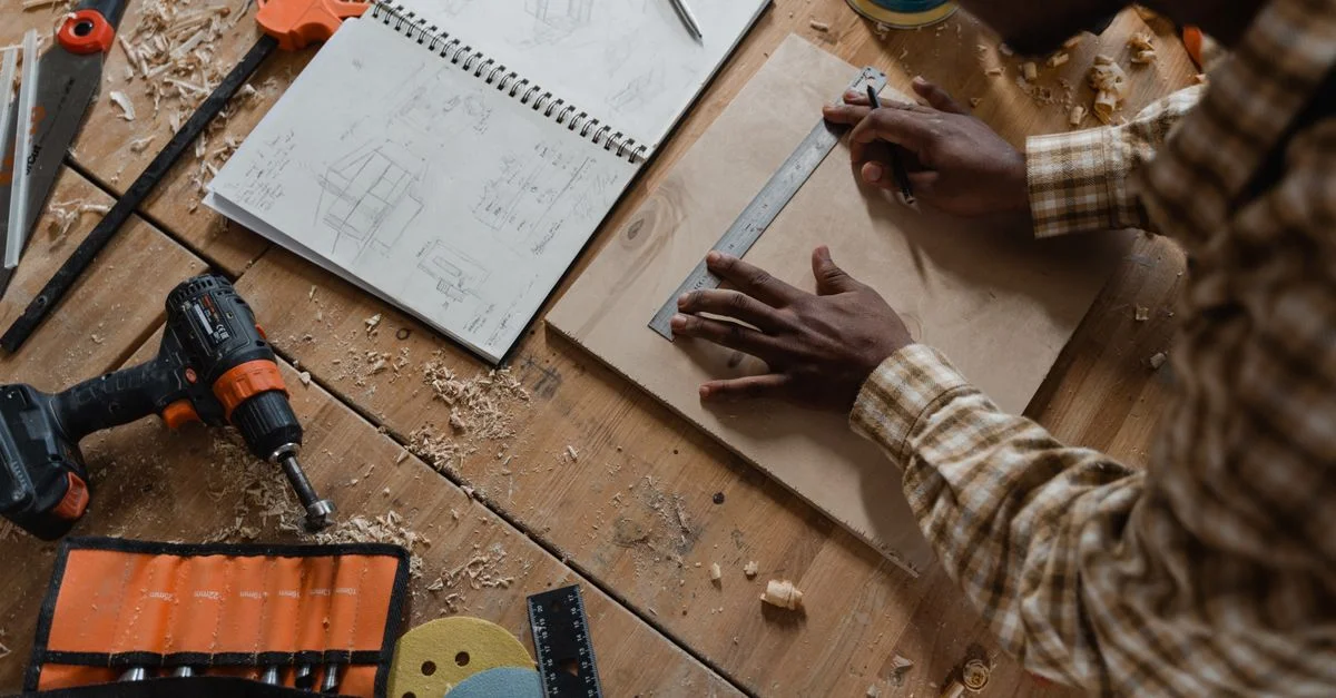 Un menuisier mesure du bois parmi des outils et des croquis dans un atelier détaillé.