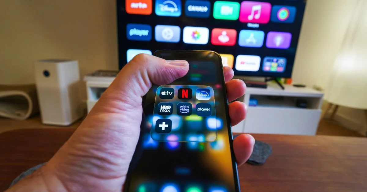 Gros plan sur une main tenant un téléphone affichant des applications de streaming devant un téléviseur comportant de multiples icônes d'applications.