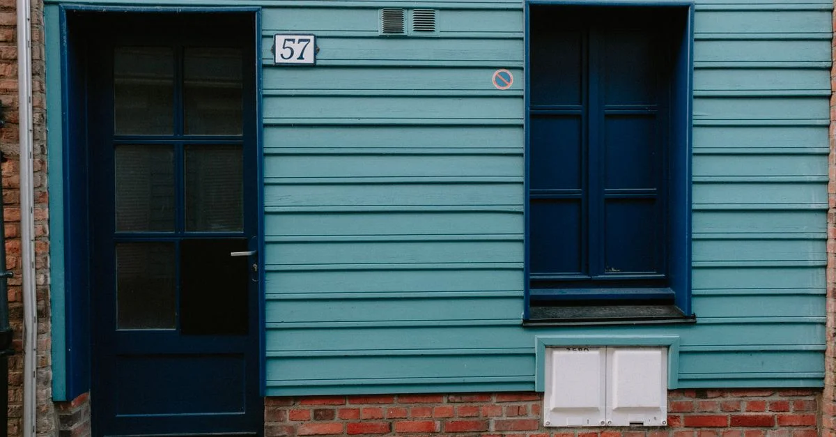 Gros plan d'une façade de bâtiment turquoise avec une porte et une fenêtre bleues, présentant une fondation en briques.