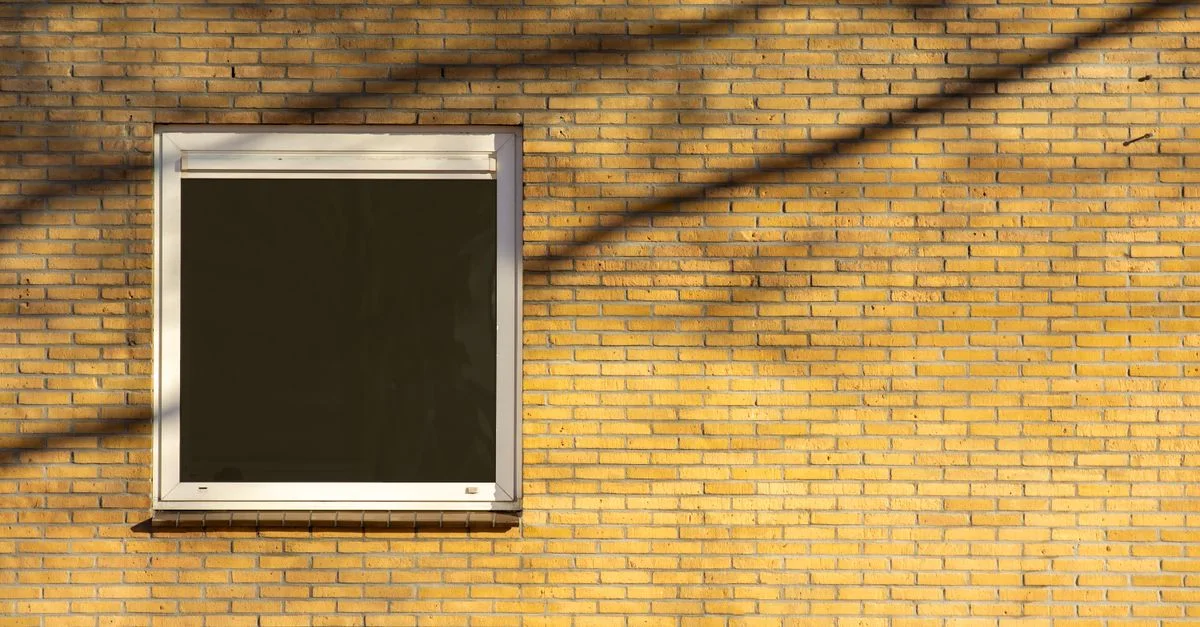 Une scène urbaine minimaliste présentant une seule fenêtre dans un mur en brique jaune, mise en valeur par des ombres.