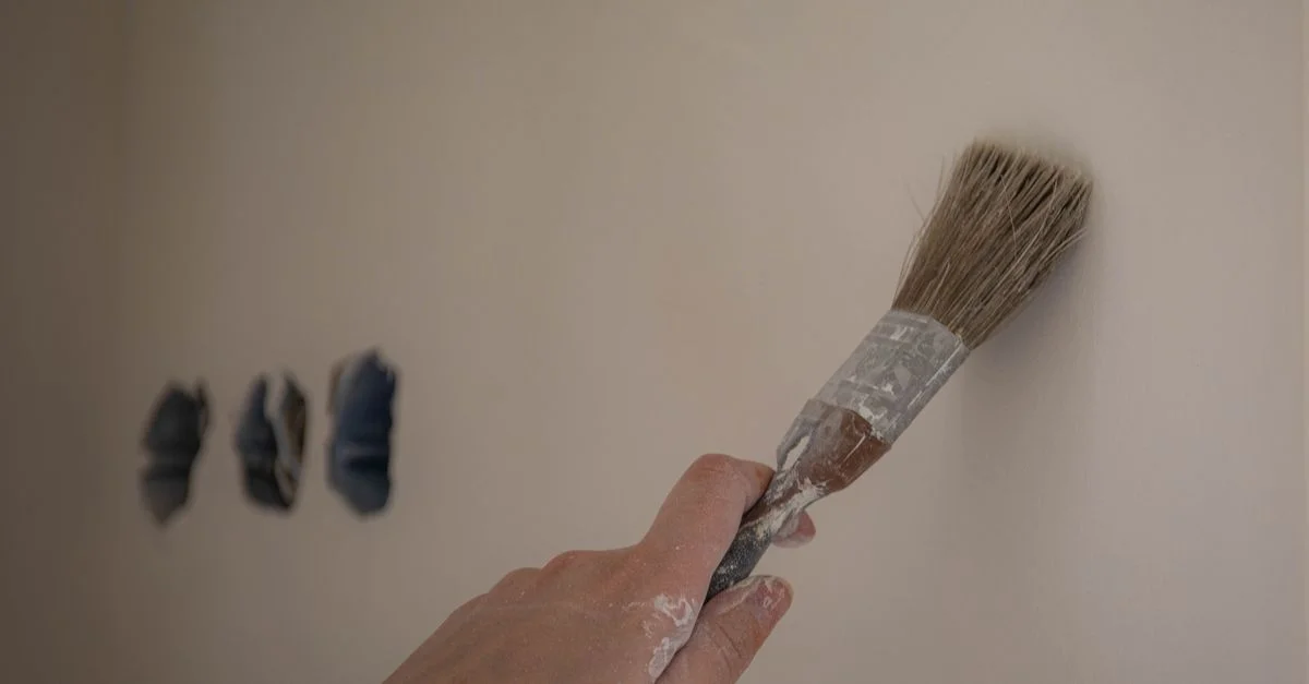 Un gros plan d'une main utilisant un pinceau pour peindre un mur blanc, trois échantillons de couleur visibles.