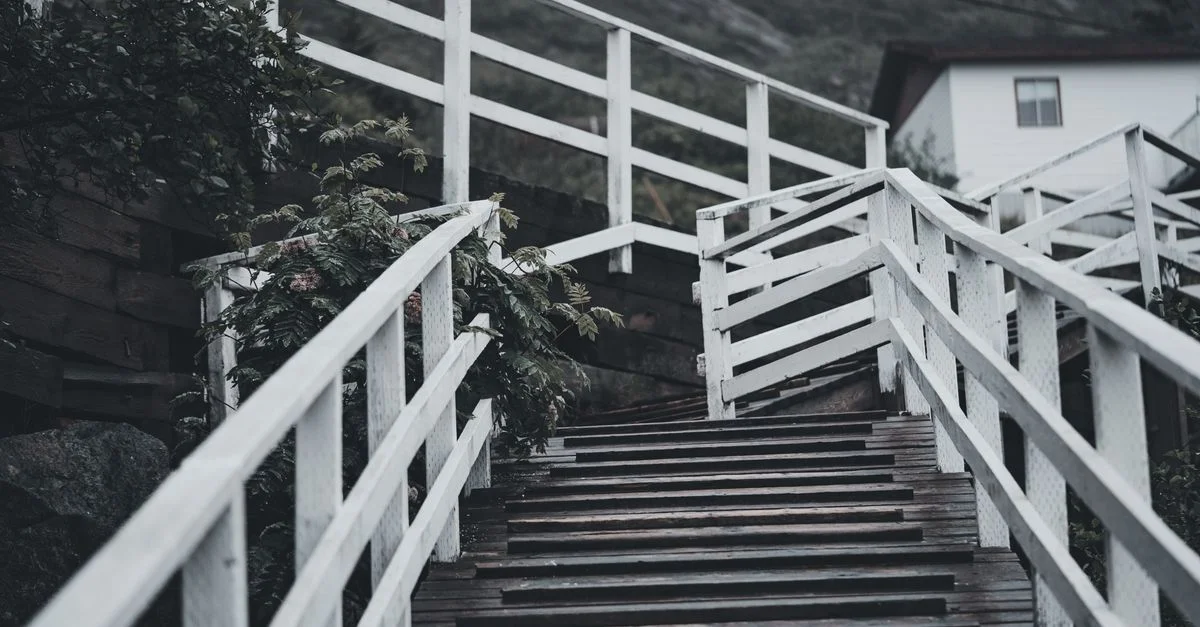 Escalier en bois rustique avec des rampes blanches menant à une maison, entourée de verdure luxuriante.
